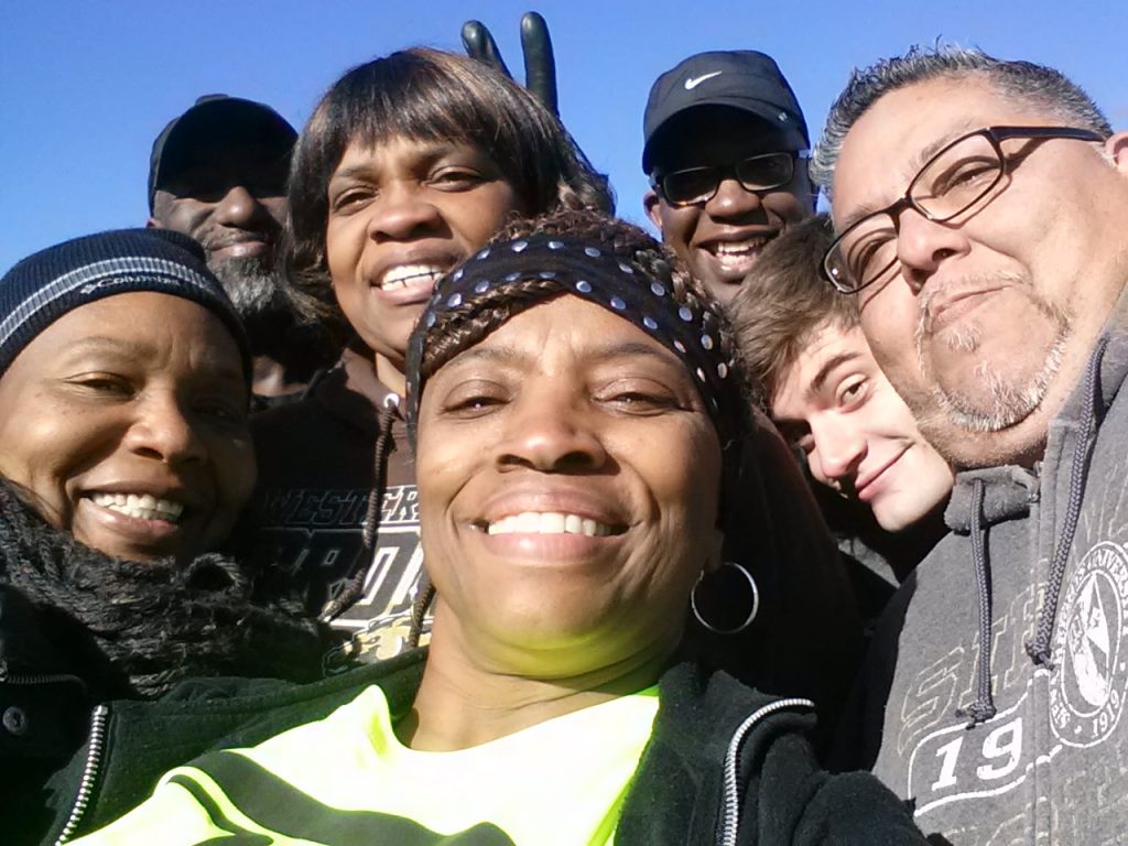 A group of smiling people pose closely together for a selfie outdoors on a sunny day.