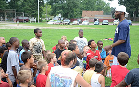A coach talks to a group of kids gathered on a grassy field during a daytime outdoor event.