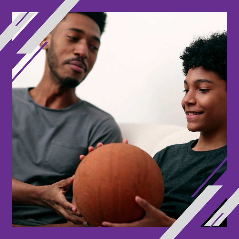 Two people sit together, smiling as they hold a basketball inside a purple and white geometric border.