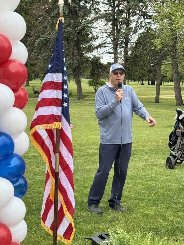 Man speaking into microphone next to U.S. flag and balloon column at outdoor event on a grassy lawn.