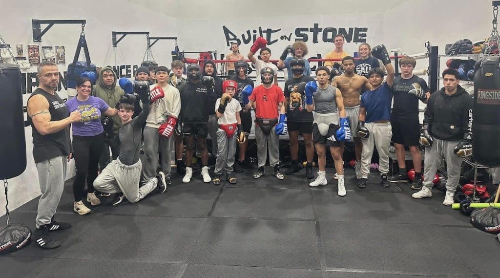 A large group of people in boxing gear pose together inside a gym with "Built on Stone" written on the wall.