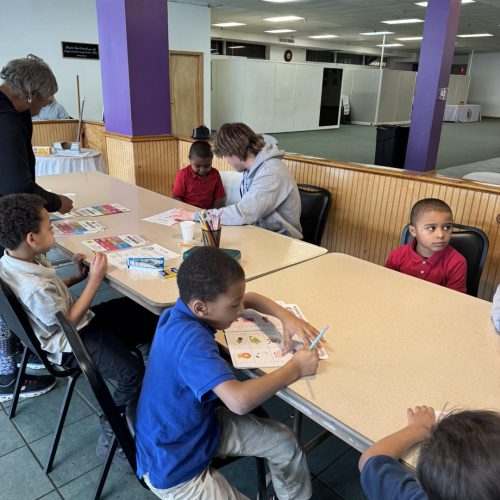 Children sit around a table coloring and doing activities, supervised by two adults in a classroom setting.